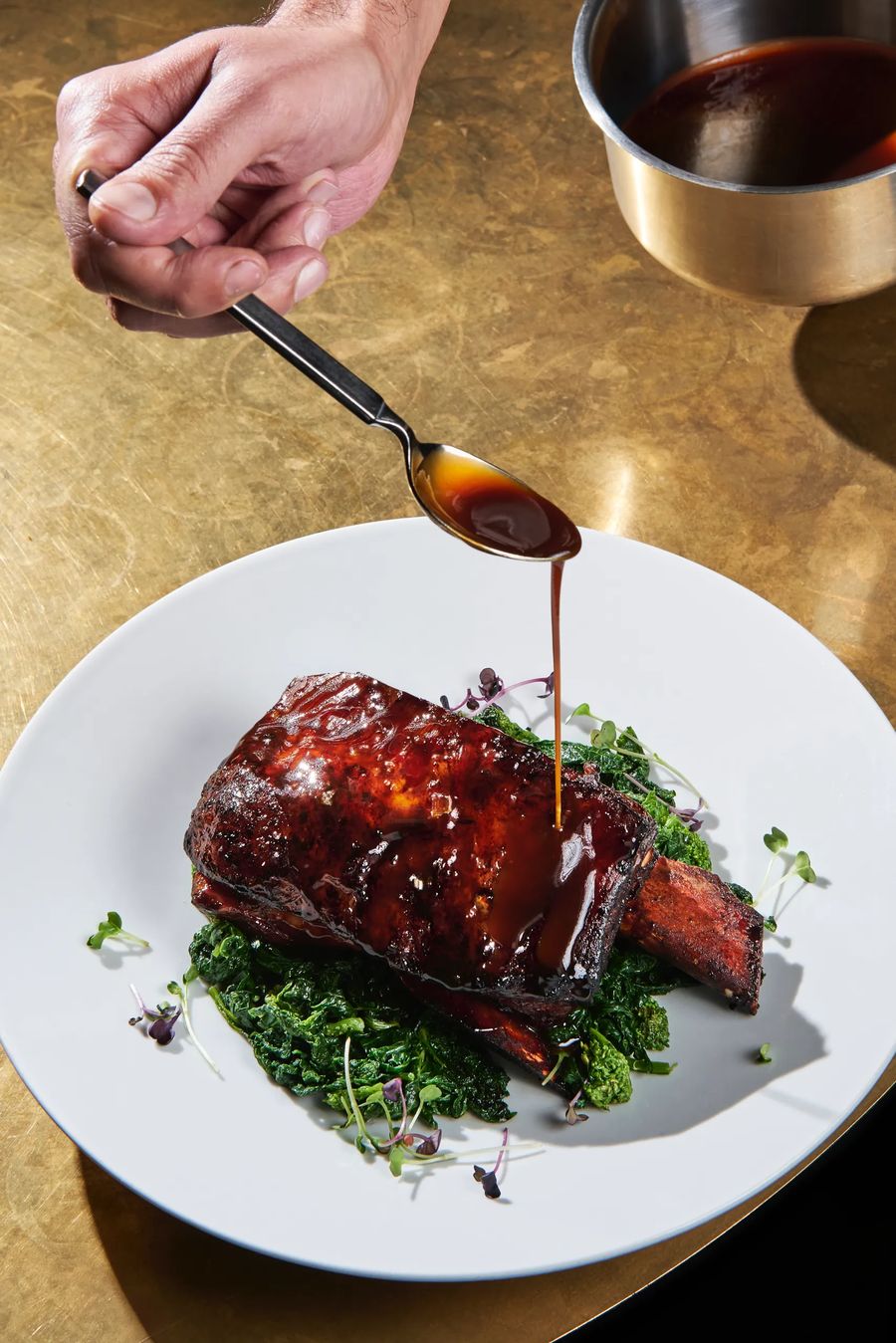 Chef che versa salsa lucida su costolette di manzo caramellate con rapini su fondo dorato — fotografo cucina gourmet Potafiori Milano
