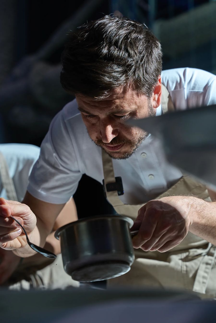 Chef in grembiule che assaggia dalla pentola nera in cucina — reportage fotografico backstage ristorante professionale Milano