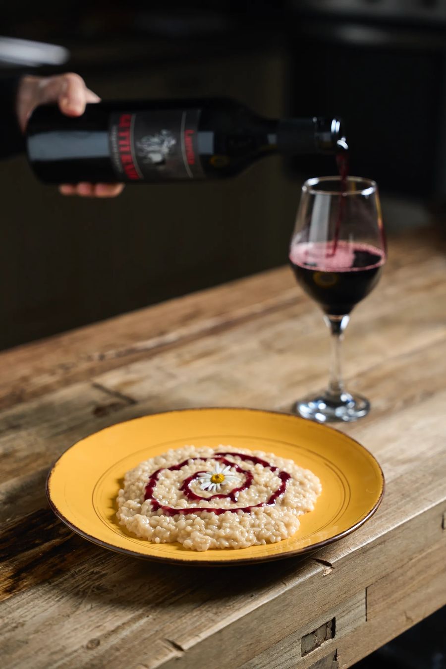 Risotto bianco con spirale di salsa rossa e margherita su piatto ocra, vino rosso versato sullo sfondo — fotografo food ristorante Lombardia