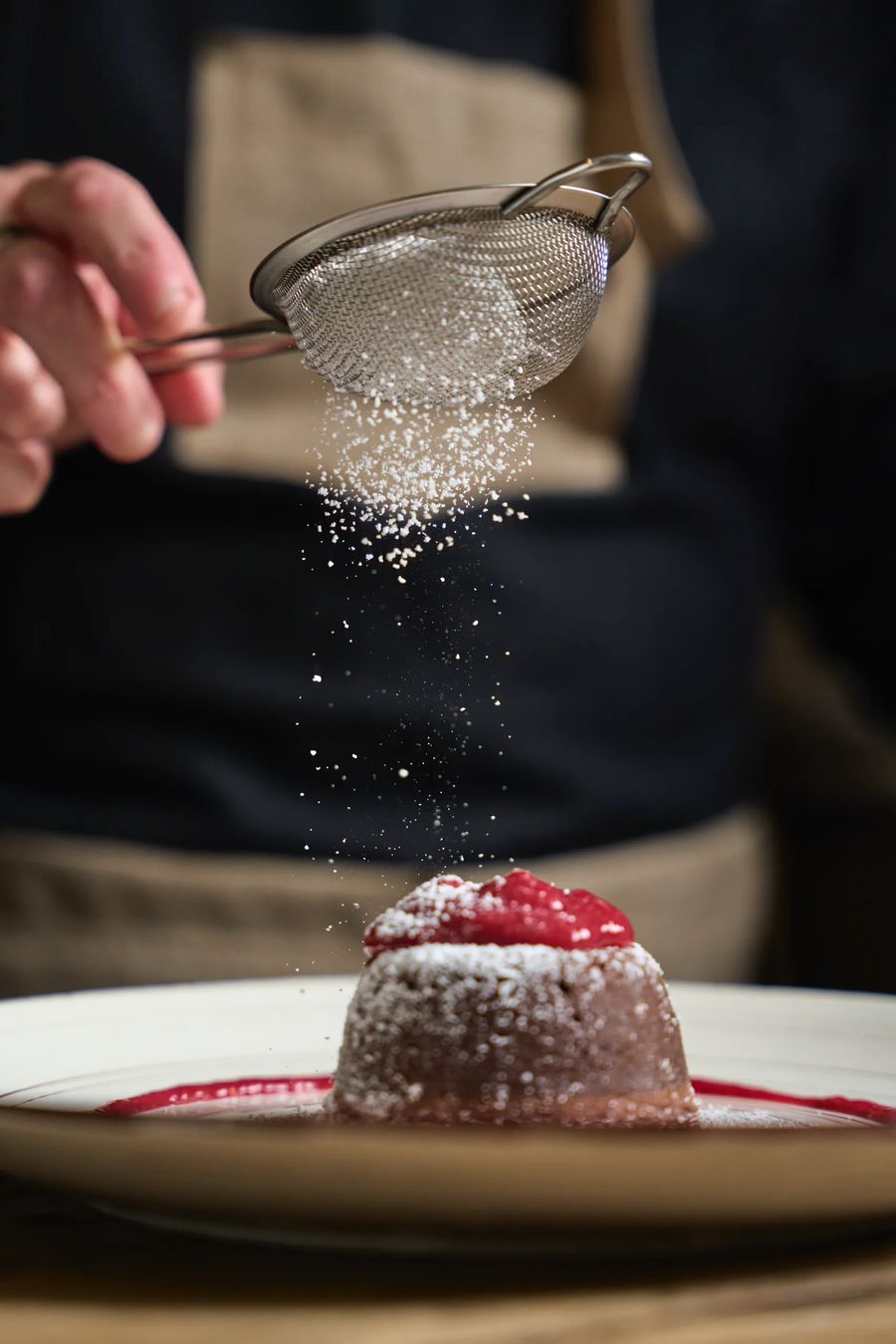 Cuoco che setaccia zucchero a velo su tortino al cioccolato con coulis di lamponi — azione pasticceria, fotografo dessert ristorante Milano