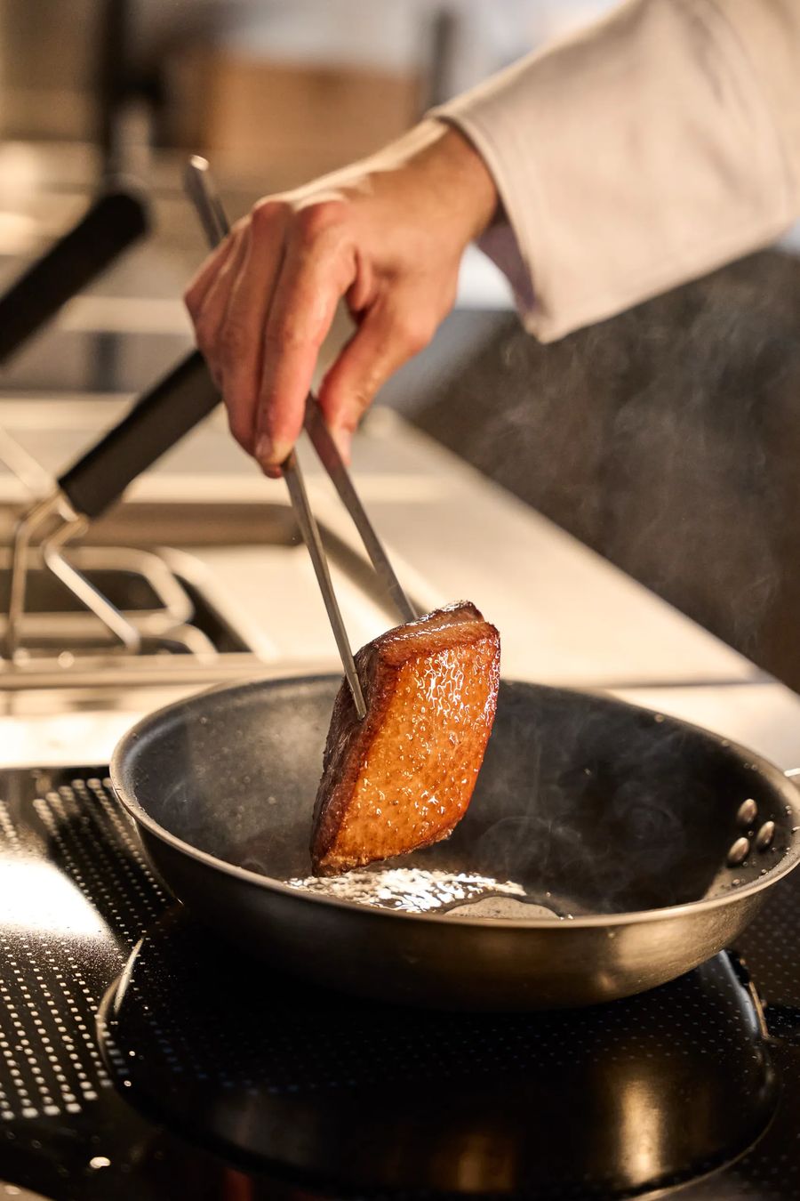 Mano del cuoco che rigira cipollotti e scalogni alla brace viva — backstage cucina, fotografo reportage ristorante Milano Lombardia