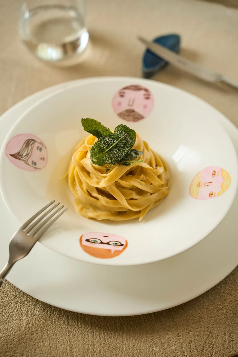Nido di tagliatelle al burro e salvia su piatto illustrato con volti colorati — pasta all'uovo, fotografia gastronomica ristorante FATA Milano