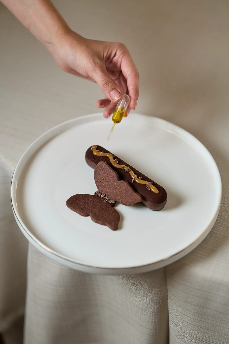 Dessert al cioccolato a forma di farfalla con caramello e granella di cacao, mano con pipetta d'olio — fotografia pasticceria gourmet Milano