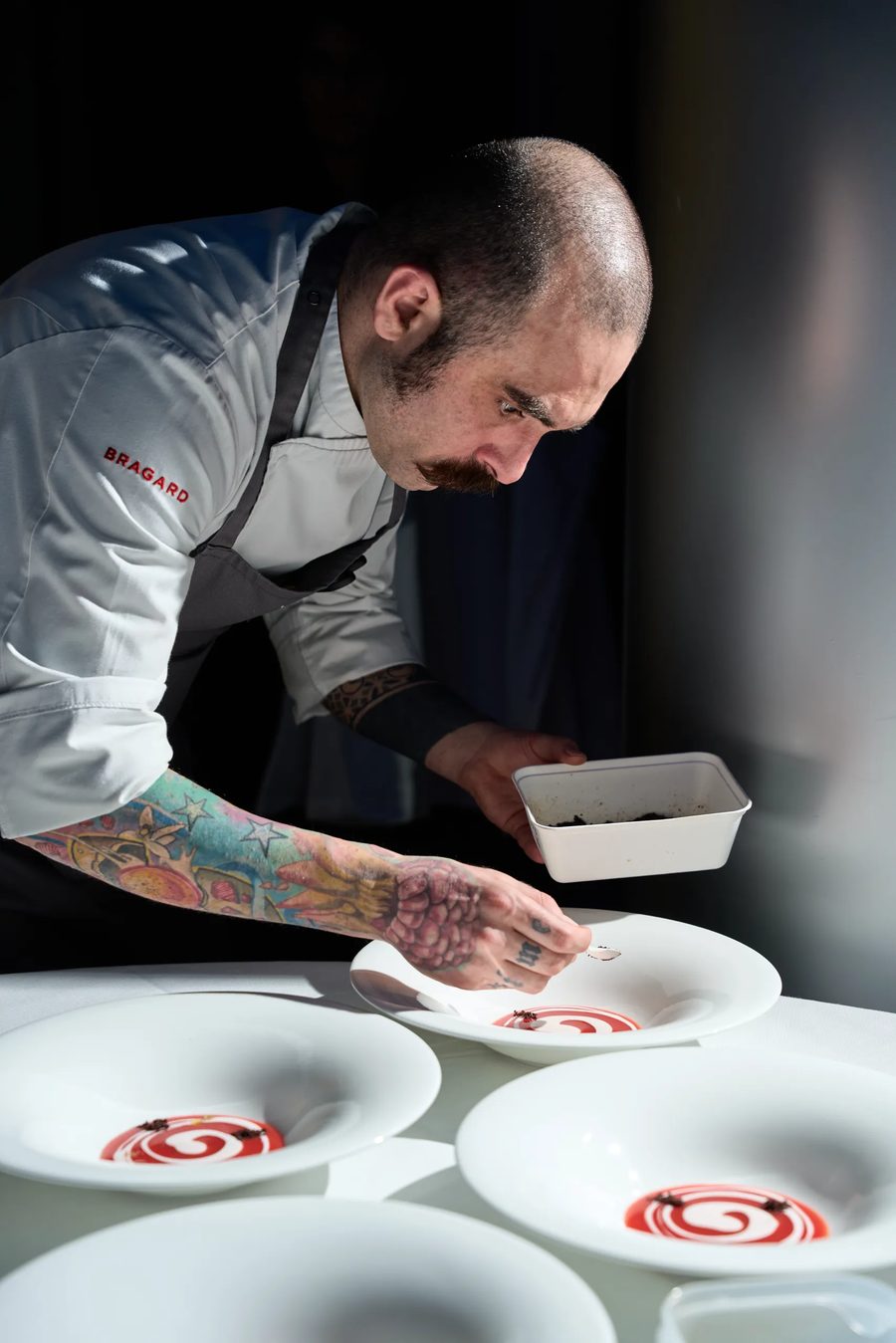 Chef con tatuaggi che impatta piatti con spirale di salsa rossa — reportage backstage cucina professionale Milano